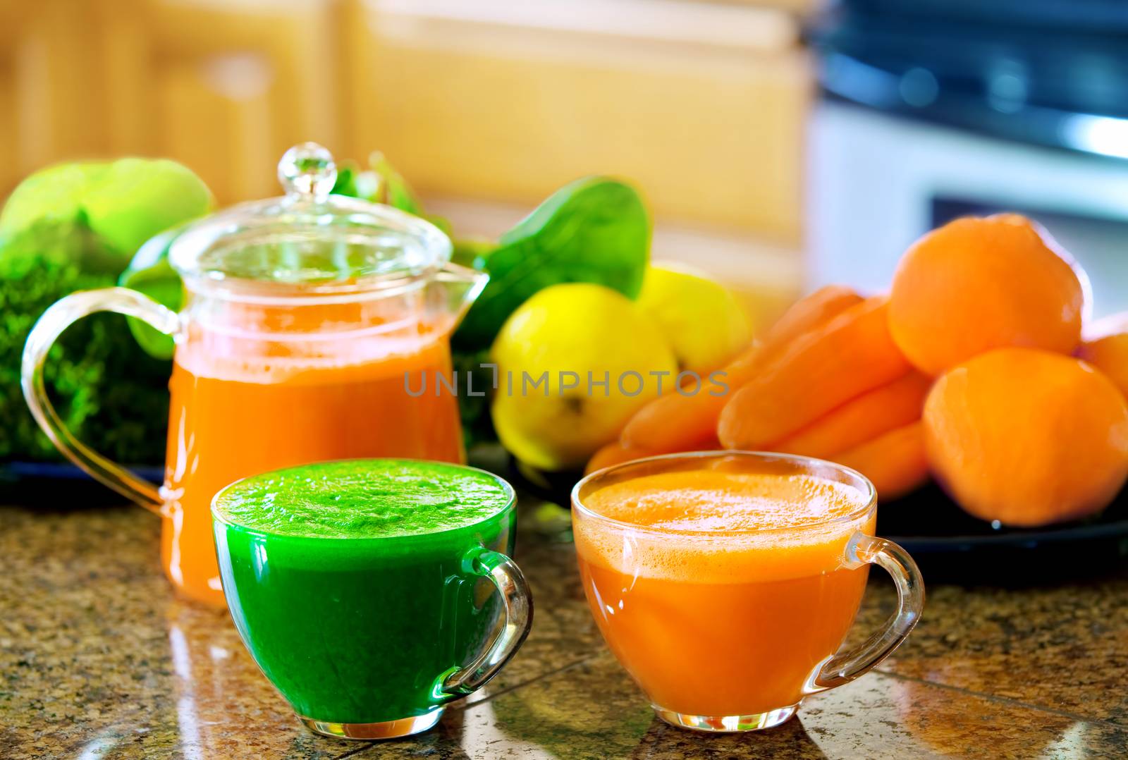 Two cups of fresh vegetable juice on kitchen counter with vegetables in background. Kale, spinach, cucumber, carrots, apples, lemons, lime, oranges.