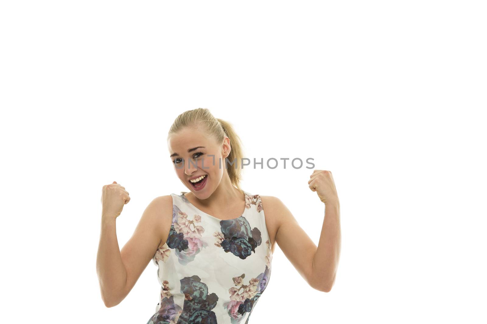 Exuberant young blond woman in a floral summer dress standing cheering and punching the air in her excitement at a success or victory, isolated on white