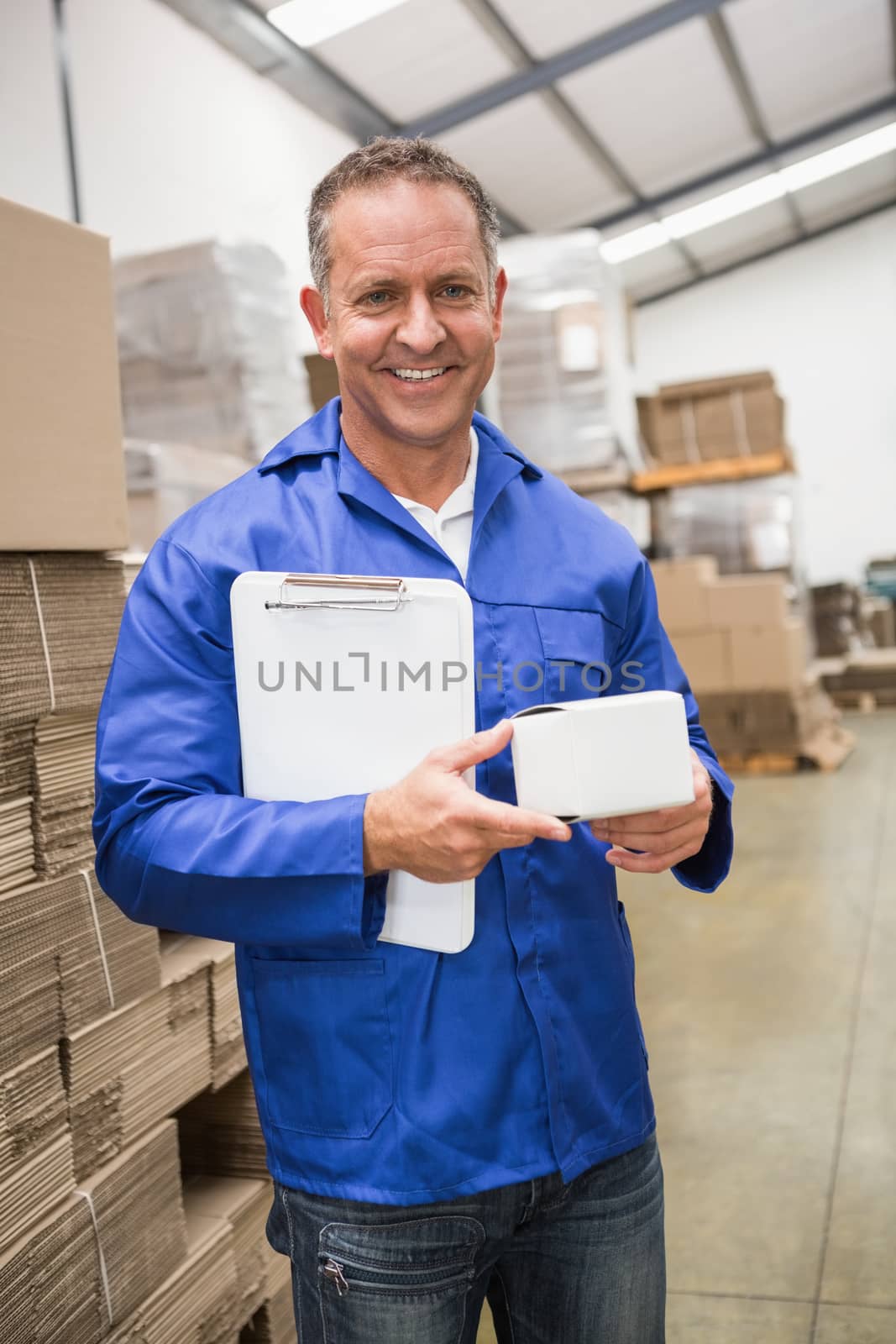Smiling warehouse worker holding small box and clipboard in a large warehouse
