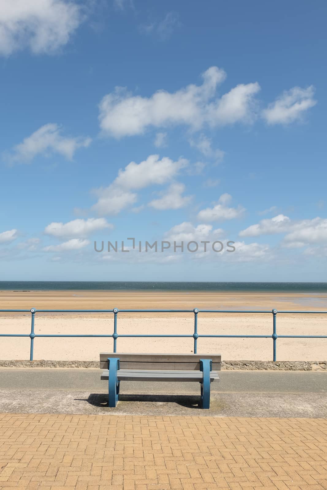 A blue painted bench and blue railings with a view across a deserted sand beach with the sea and blue cloudy sky in the distance.