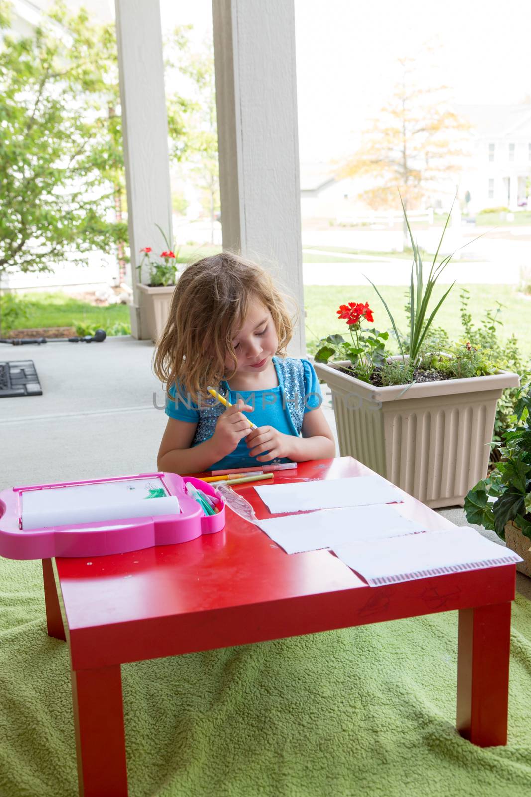 Cute little girl kneeling down at a colorful vivid red table coloring in with pencil crayons on an outdoor undercover patio in spring or summer