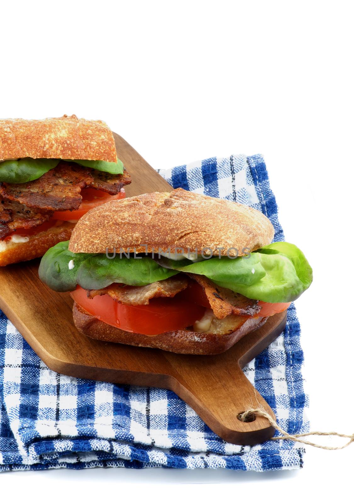 Delicious Ciabatta Sandwiches with Bacon, Tomato, Lettuce and Sauces on Cutting Board and Checkered Napkin isolated on white background