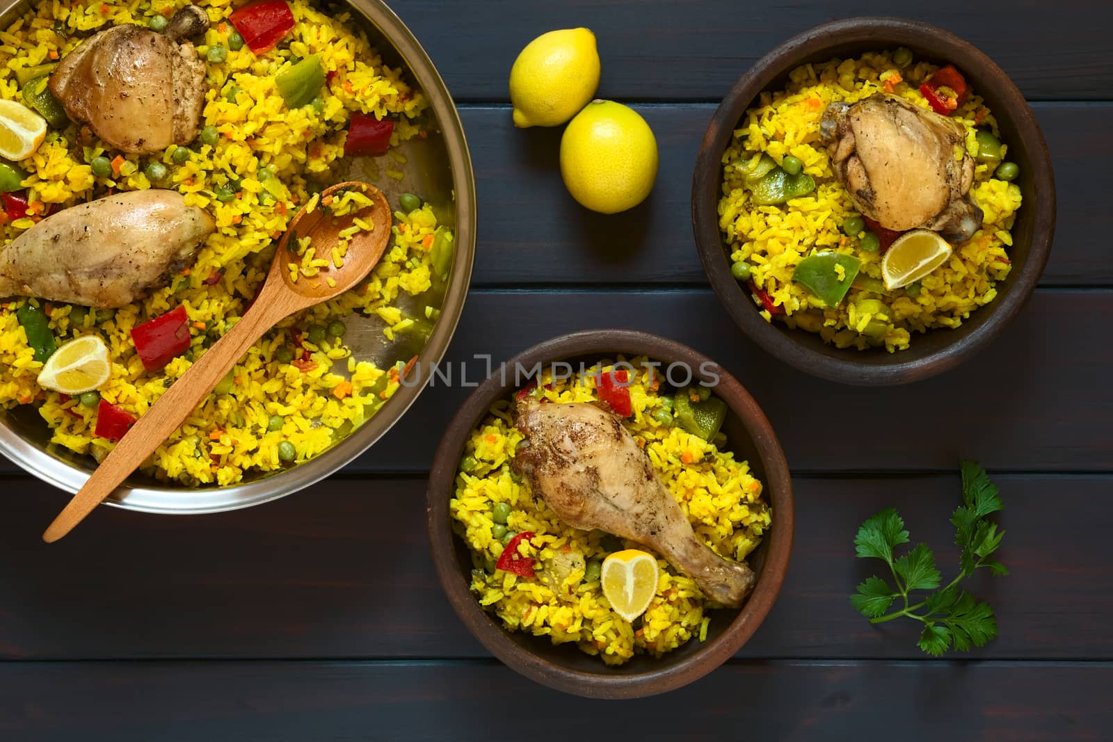 Overhead shot of two rustic bowls and a pot of chicken paella, a traditional Valencian (Spanish) rice dish made of rice, chicken, peas and capsicum and served with lemon, photographed on dark wood with natural light