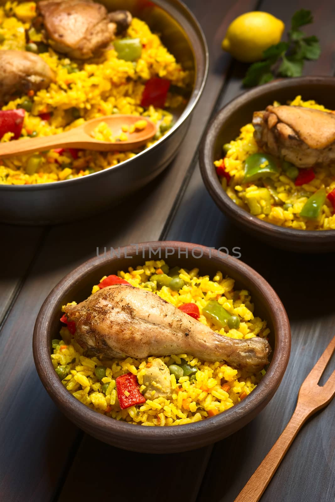 Two rustic bowls and a pot of chicken paella, a traditional Valencian (Spanish) rice dish made of rice, chicken, peas and capsicum , photographed on dark wood with natural light (Selective Focus, Focus on the chicken thigh)
