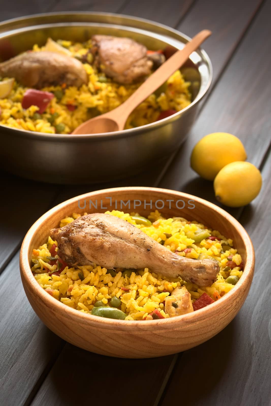 Wooden bowl and a pot of chicken paella, a traditional Valencian (Spanish) rice dish made of rice, chicken, peas and capsicum , photographed on dark wood with natural light (Selective Focus, Focus on the chicken thigh)