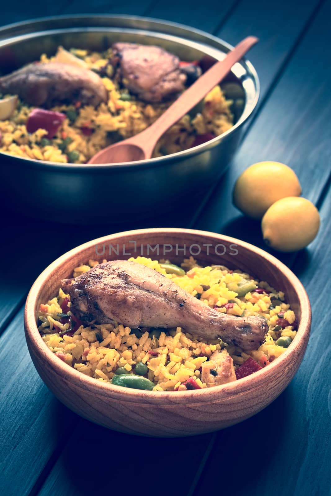 Toned image of a wooden bowl and a pot of chicken paella, a traditional Valencian (Spanish) rice dish made of rice, chicken, peas and capsicum , photographed on dark wood with natural light (Selective Focus, Focus on the chicken thigh)