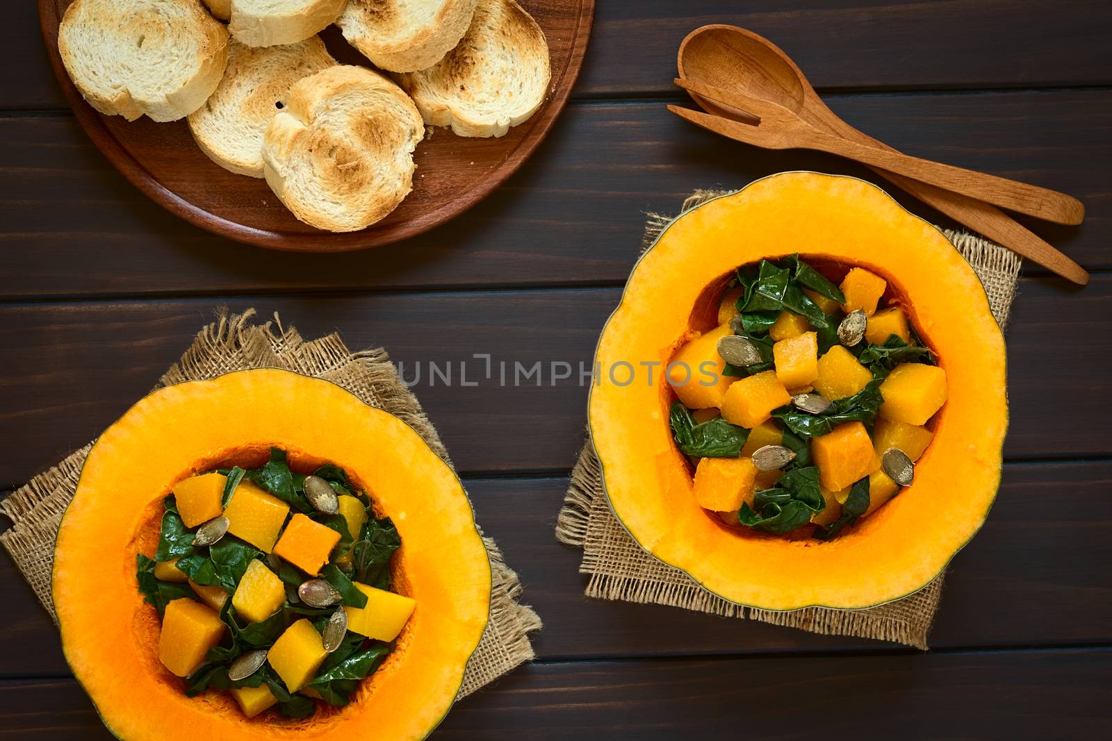 Pumpkin and chard salad with roasted pumpkin seeds served in pumpkin halves with toasted bread on the side, photographed overhead on dark wood with natural light  