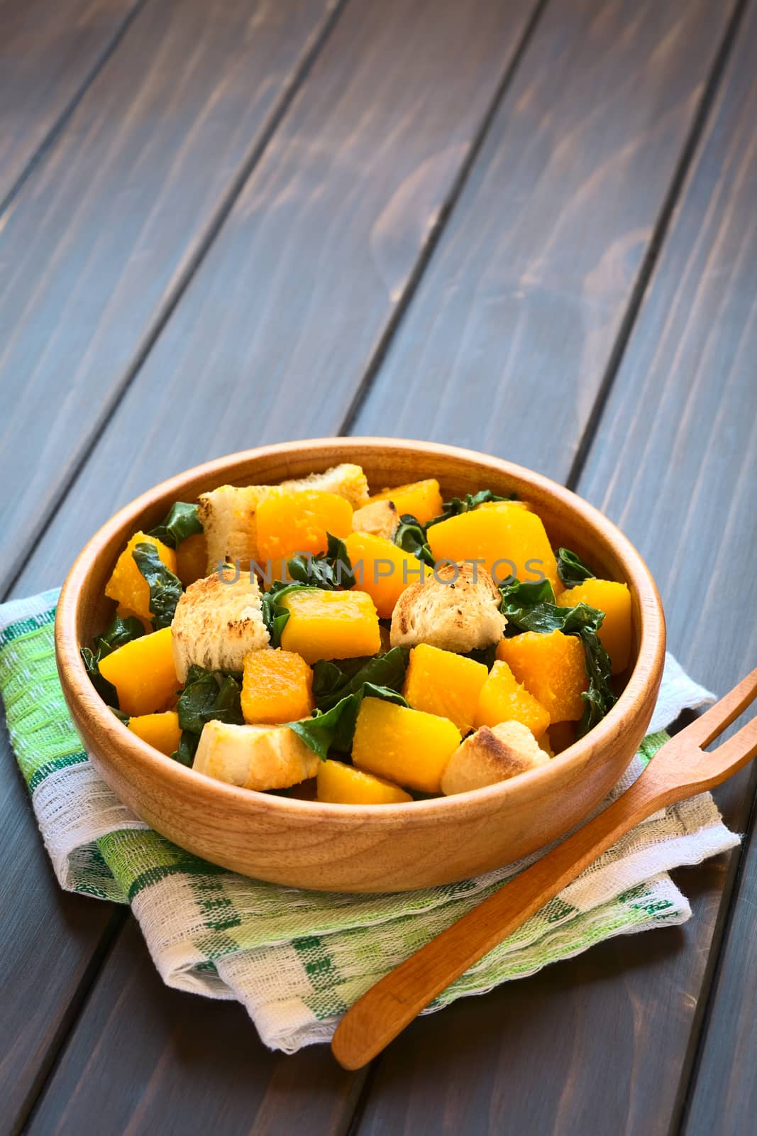 Pumpkin and chard salad with croutons served in wooden bowl, photographed on dark wood with natural light (Selective Focus, Focus in the middle of the salad) 