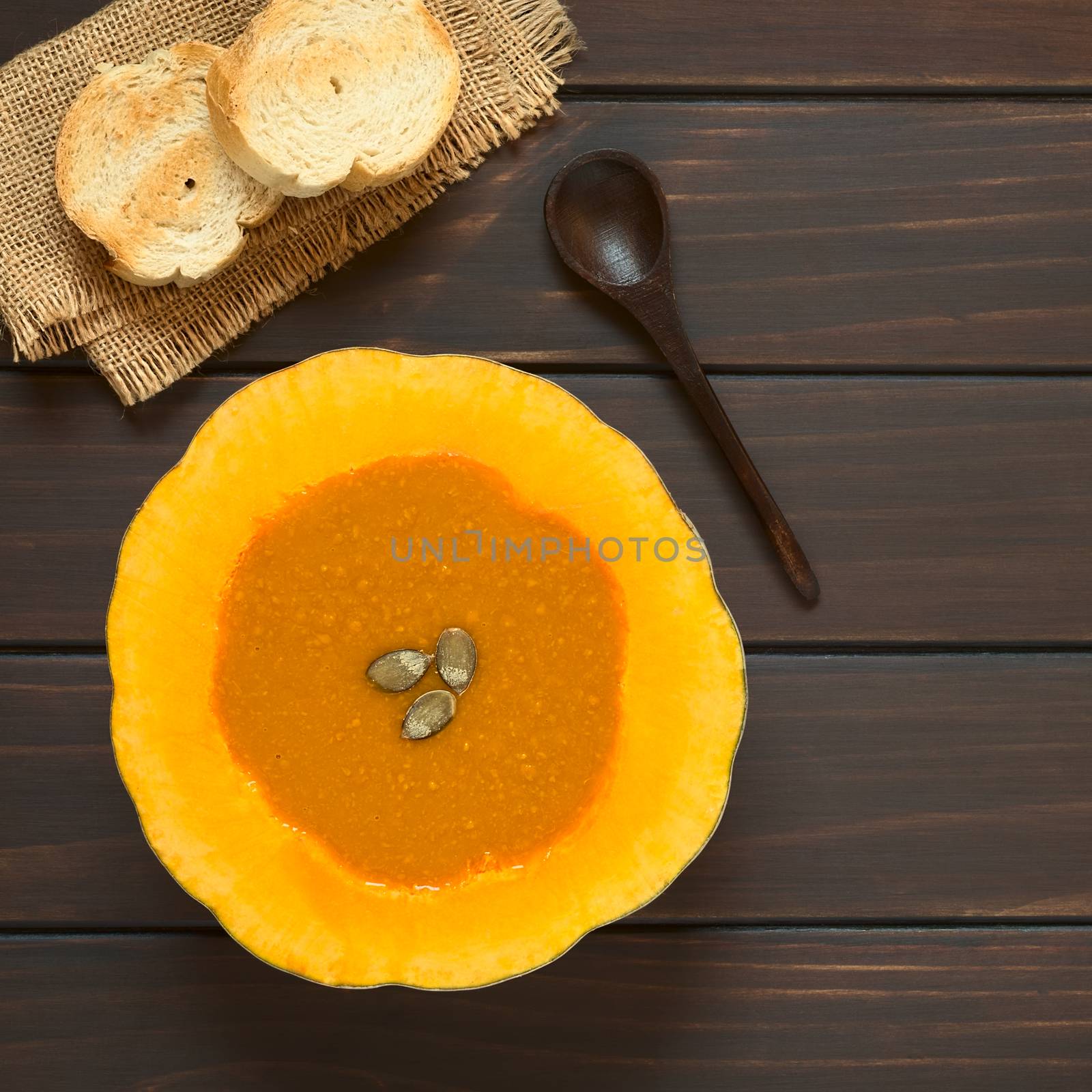 Cream of pumpkin soup served in half pumpkins with pepita seeds on top, toasted bread slices on the side, photographed overhead on dark wood with natural light