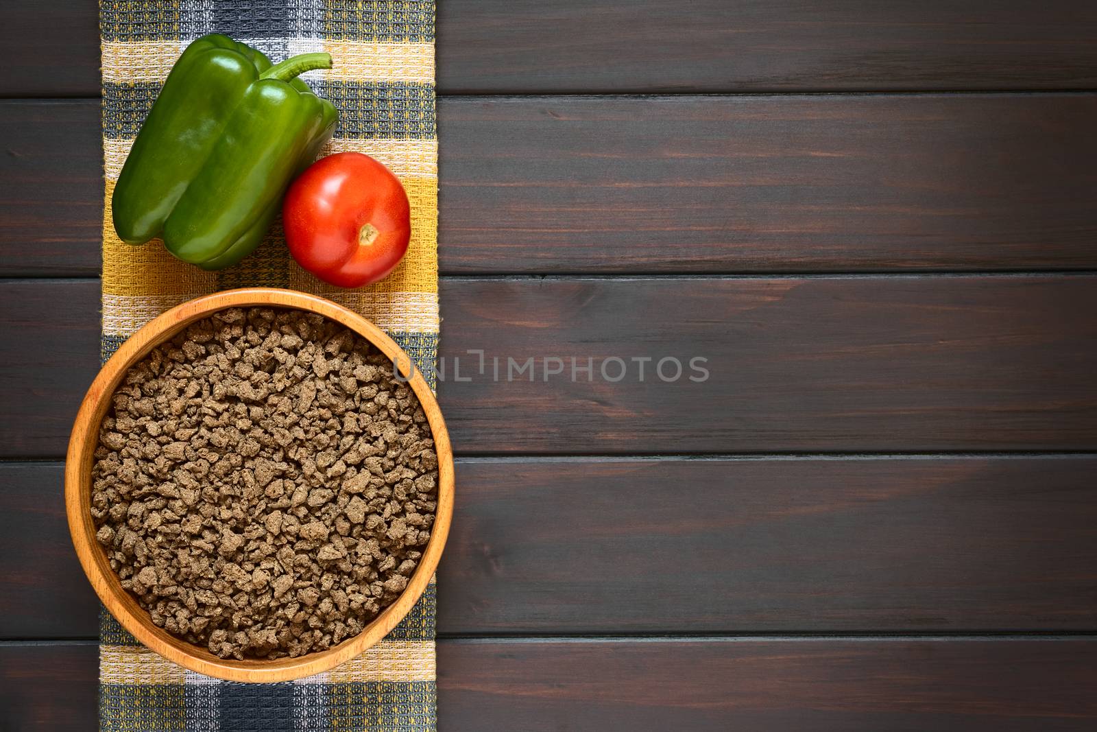 Raw textured vegetable or soy protein, called also soy meat in wooden bowl with bell pepper and tomato on kitchen towel. Photographed overhead on dark wood with natural light. 