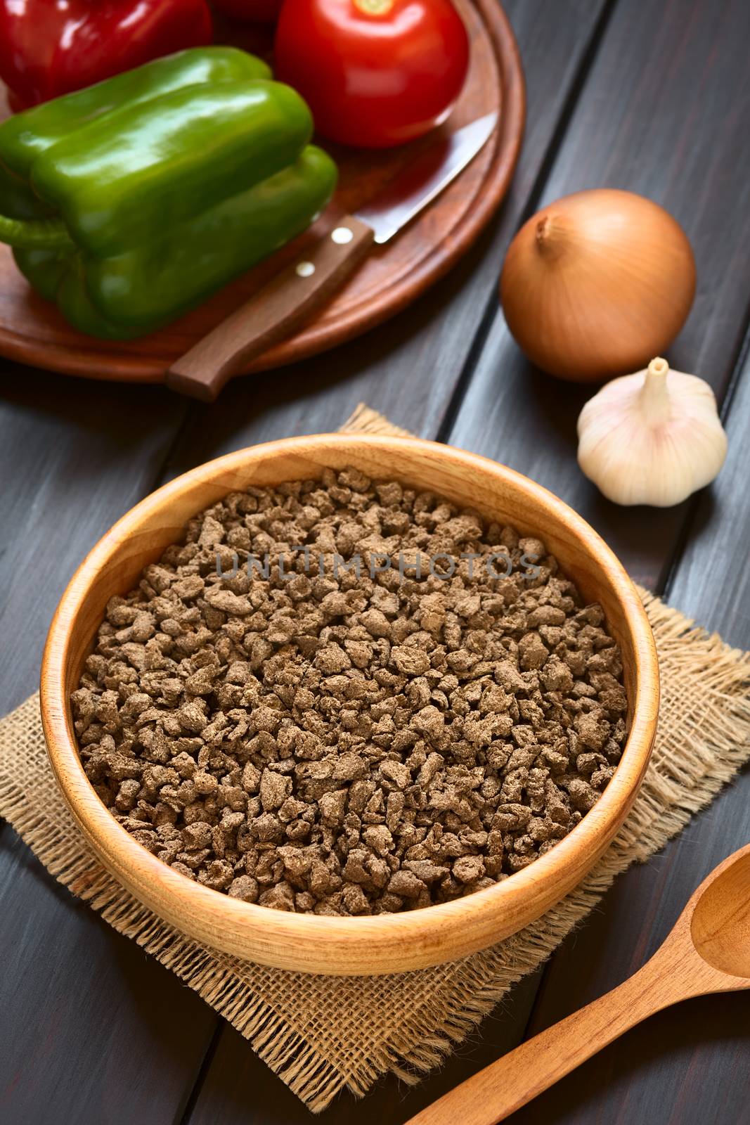 Raw textured vegetable or soy protein, called also soy meat in wooden bowl with raw vegetables in the back. Photographed on dark wood with natural light (Selective Focus, Focus one third into the soy meat) 