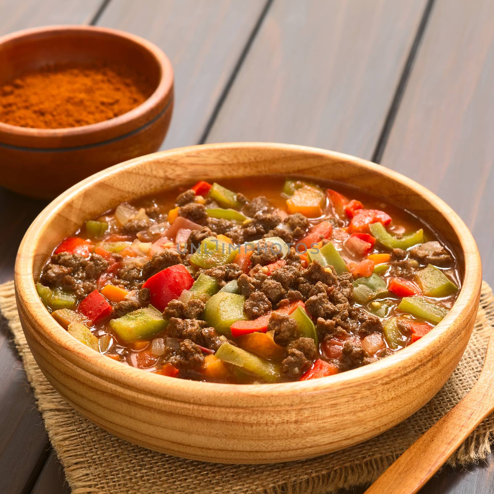 Vegan goulash made of soy meat (textured vegetable protein), capsicum, tomato and onion, served in wooden bowl, paprika powder in the back, photographed with natural light (Selective Focus, Focus one third into the dish)