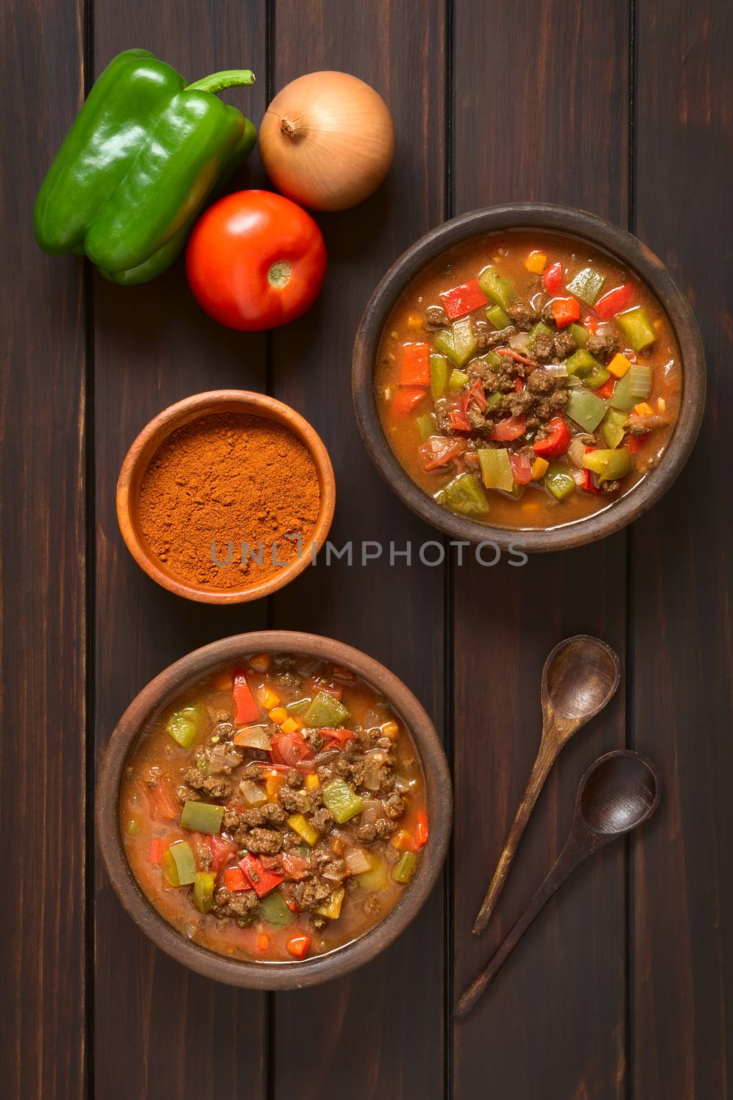 Vegan goulash made of soy meat (textured vegetable protein), capsicum, tomato and onion served in rustic bowls, paprika powder, fresh vegetables, spoons on the side, photographed overhead on dark wood with natural light