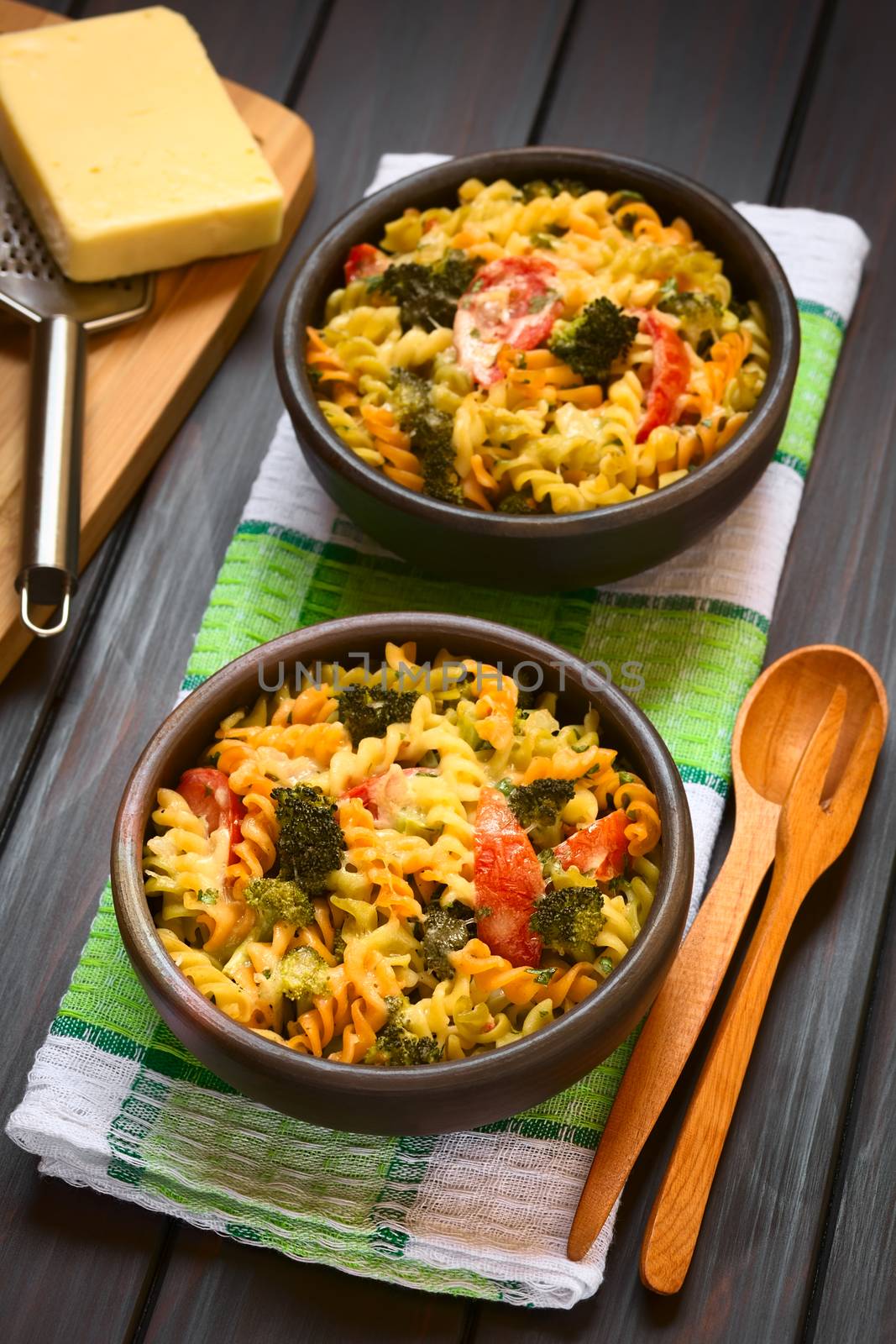 Baked tricolor fusilli pasta and vegetable (broccoli, tomato) casserole in rustic bowls, wooden cutlery on the side, grater and cheese in the back, photographed with natural light (Selective Focus, Focus in the middle of the first dish)