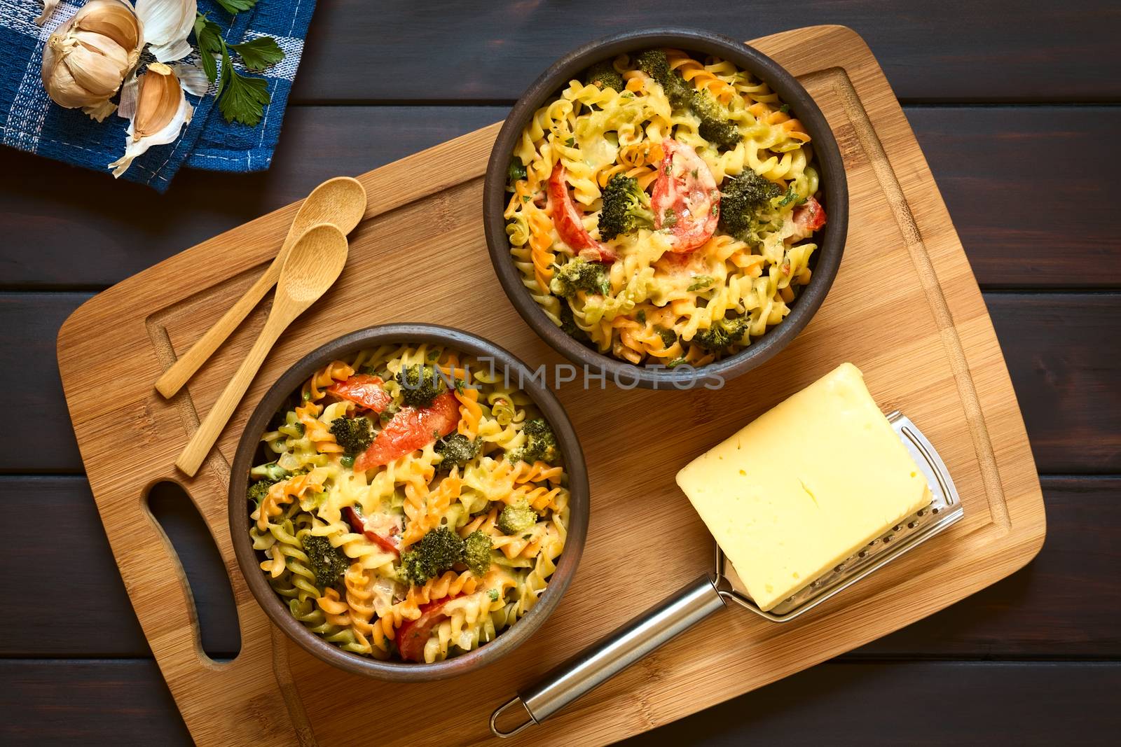 Baked tricolor fusilli pasta and vegetable (broccoli, tomato) casserole in rustic bowls, with grater, cheese and spoons on wooden board, photographed overhead on dark wood with natural light