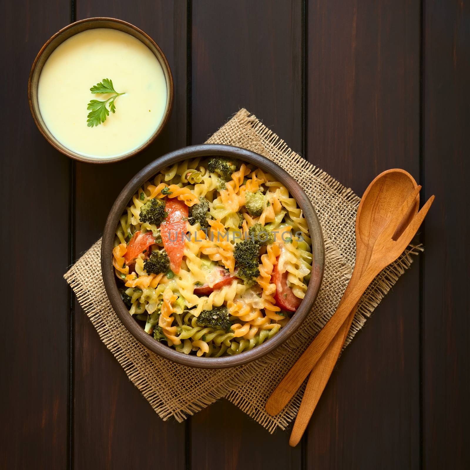 Baked tricolor fusilli pasta and vegetable (broccoli, tomato) casserole in rustic bowls, wooden spoon, fork and cream sauce on the side, photographed overhead on dark wood with natural light