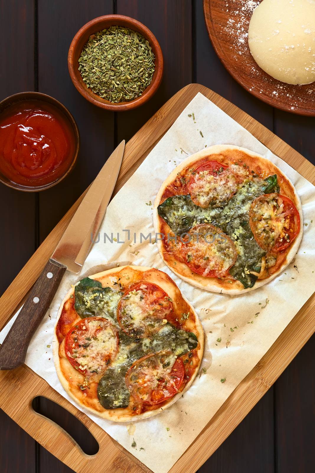 Homemade spinach and tomato pizza on baking paper on wooden board, pizza dough, dried oregano, tomato sauce on the side, photographed overhead on dark wood with natural light