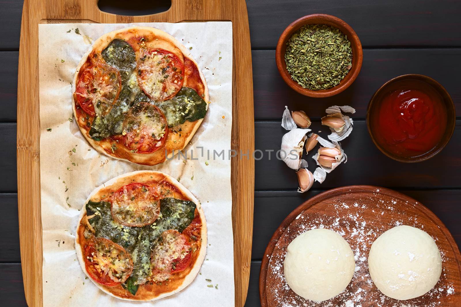 Homemade spinach and tomato pizza on baking paper on wooden board, pizza dough, dried oregano, tomato sauce, garlic on the side, photographed overhead on dark wood with natural light