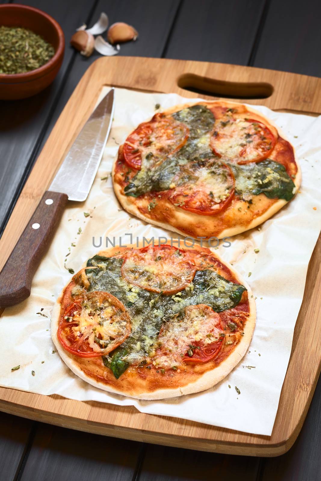Homemade spinach and tomato pizza on baking paper on wooden board, knife on the side, photographed on dark wood with natural light (Selective Focus, Focus one third into the image)