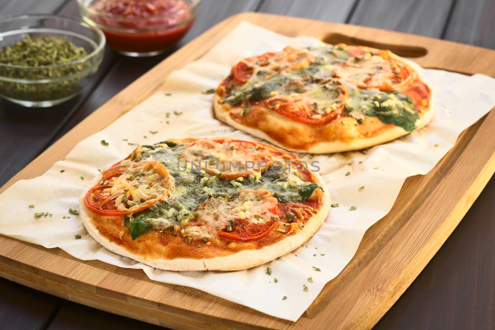 Homemade spinach and tomato pizza on baking paper on wooden board, photographed on dark wood with natural light (Selective Focus, Focus one third into the first pizza)