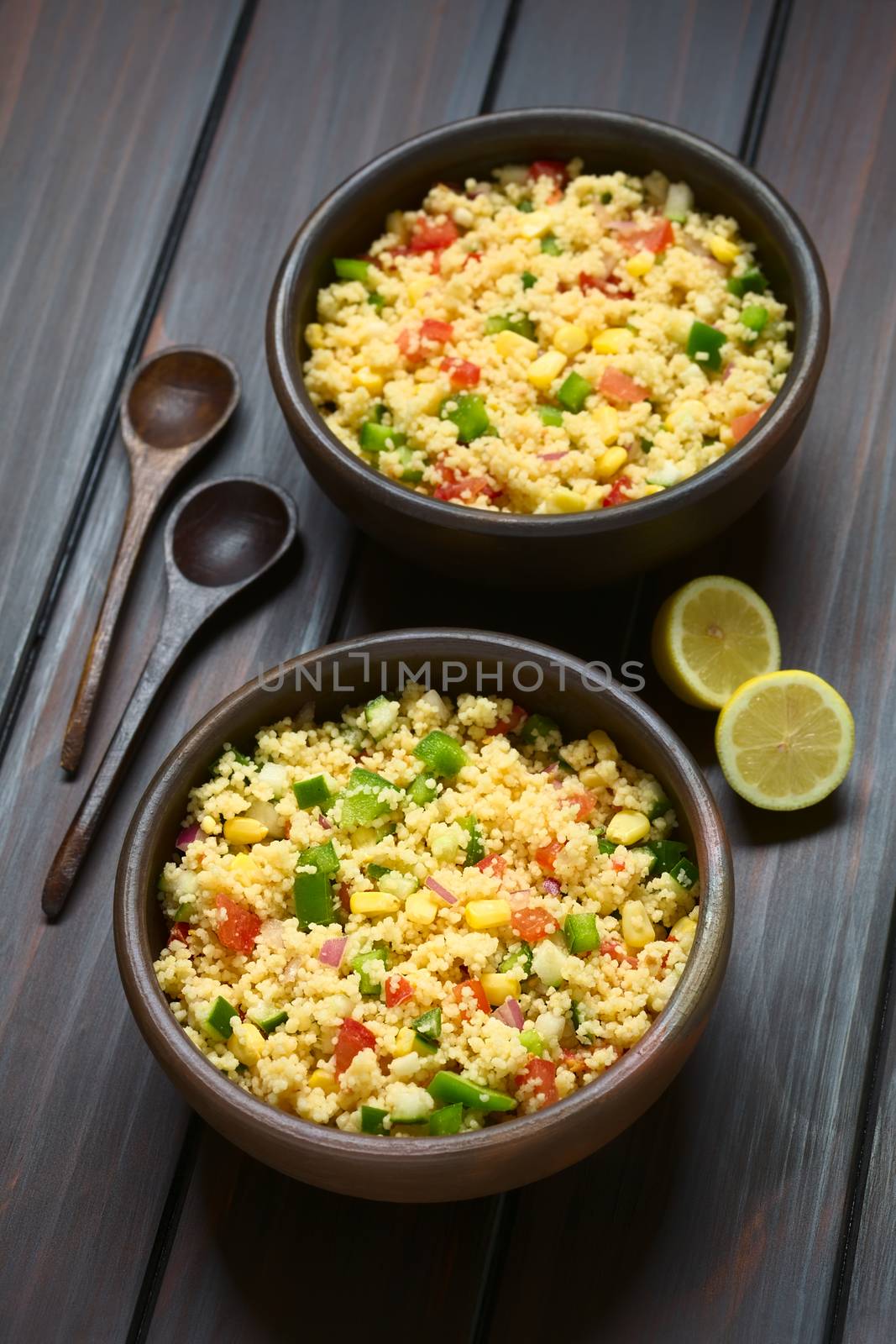 Vegetarian couscous salad made with bell pepper, tomato, cucumber, red onion and sweet corn kernels, served in rustic bowls, wooden spoons and lemon on the side. Photographed on dark wood with natural light. (Selective Focus, Focus in the middle of the first salad)