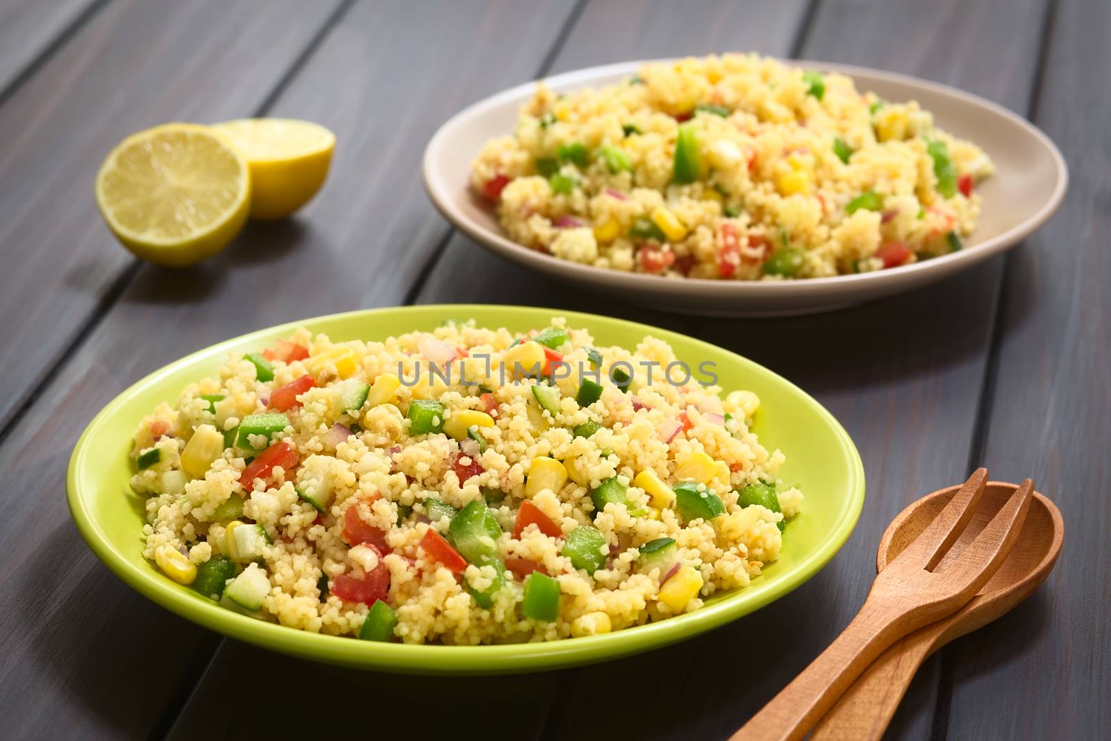 Vegetarian couscous salad made with bell pepper, tomato, cucumber, red onion and sweet corn kernels, served on plates, cutlery and lemon on the side. Photographed on dark wood with natural light. (Selective Focus, Focus one third into the first salad)