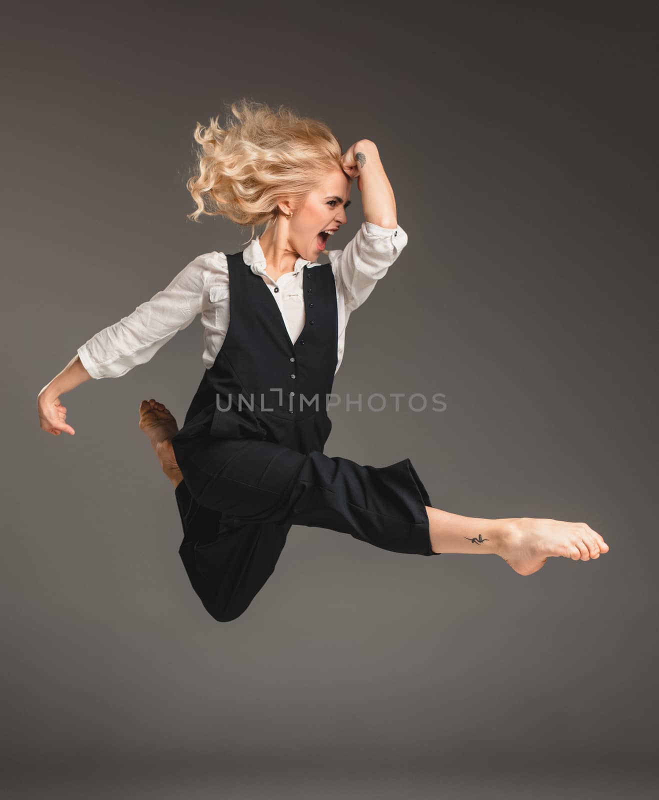 Beauty blond woman in a black suit and white shirt in ballet jump on a gray background