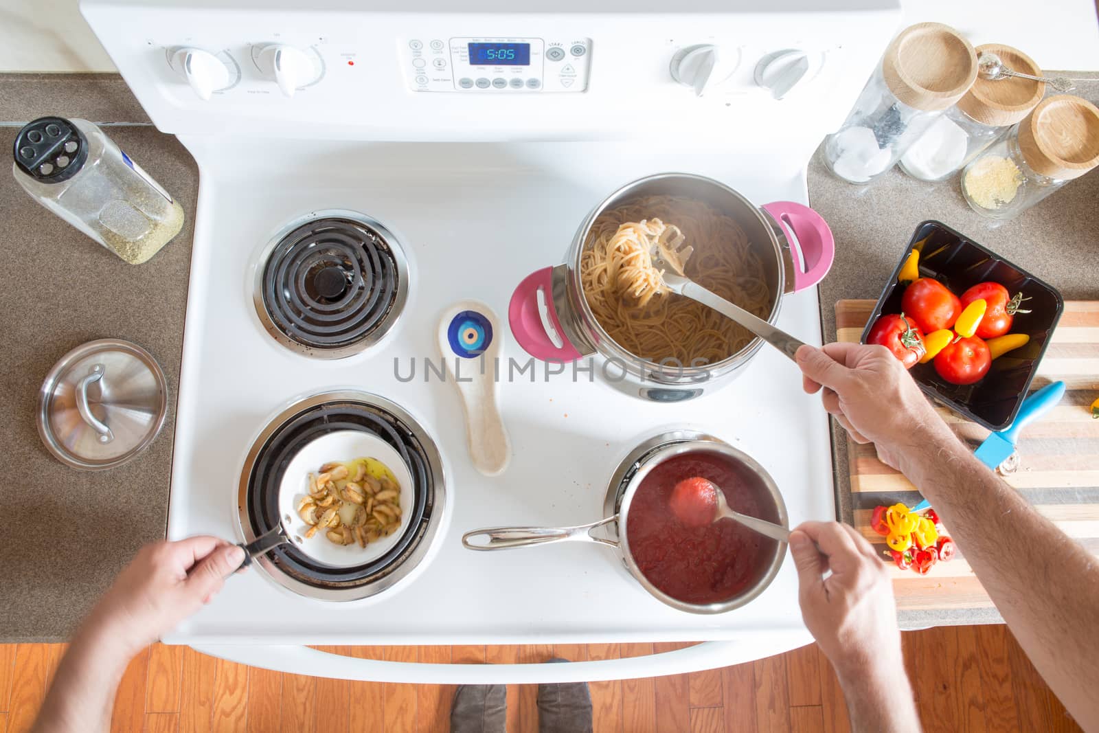 Two men preparing healthy brown wholewheat spaghetti cooking together at the hob as a team stirring the boiling pasta and sauce, overhead view of their hands and ingredients