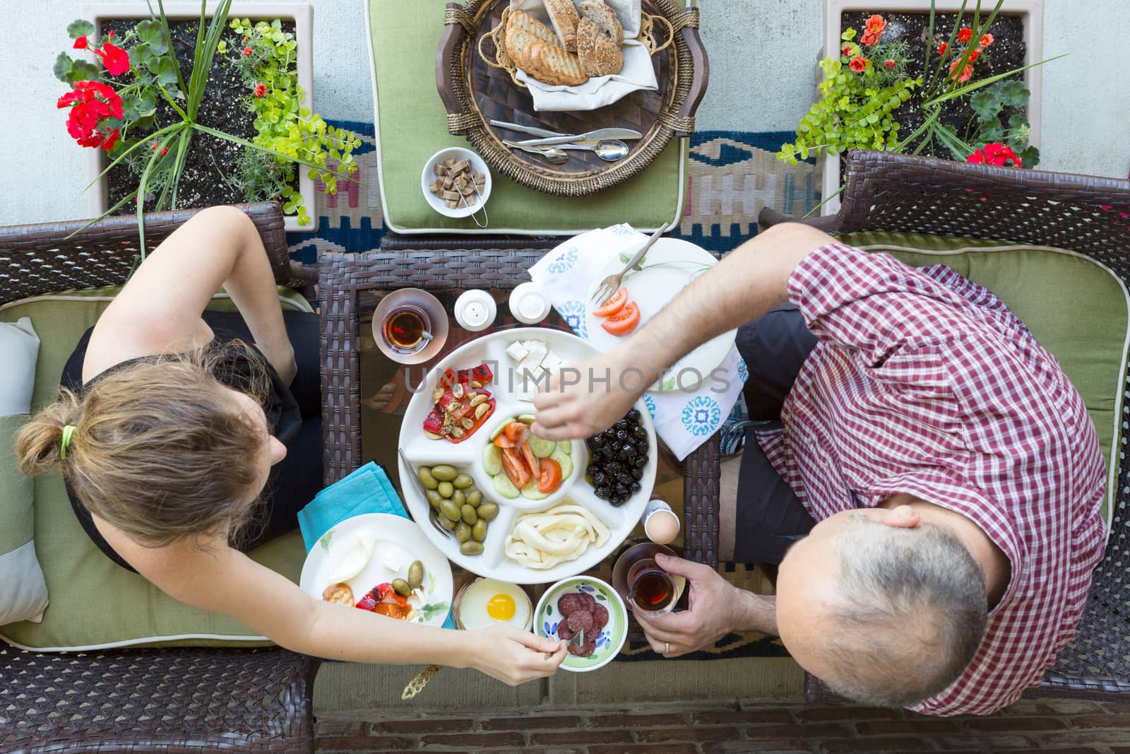 Man and woman enjoying an outdoor Turkish breakfast sitting together at a small intimate table helping themselves to a selection of food with Turkish tea, overhead view