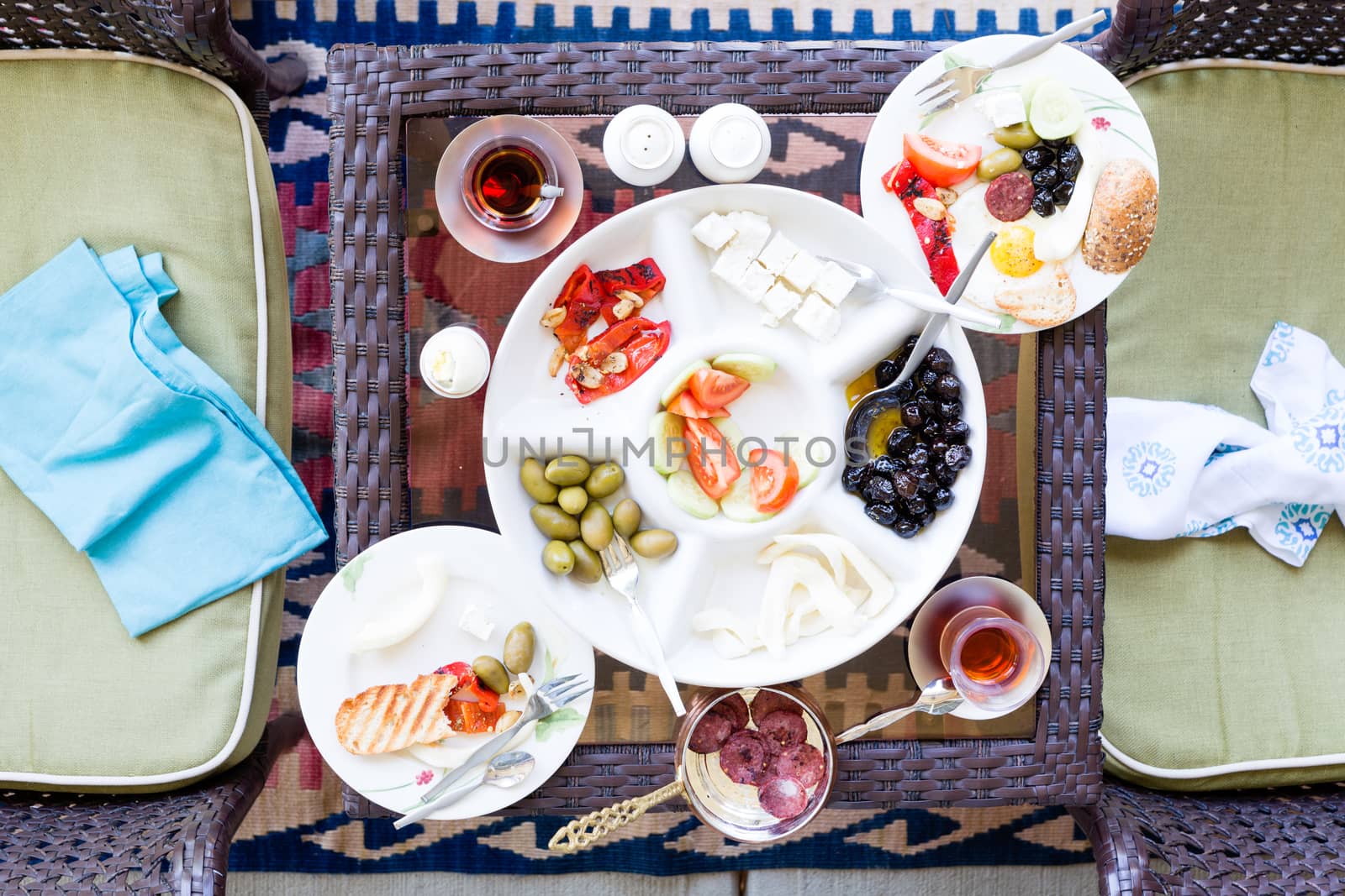 Unfinished Turkish breakfast on a patio table with a serving of fried eggs with a selection of fresh tomato, olives, cheese and mugs of Turkish tea, overhead view with napkins on chairs