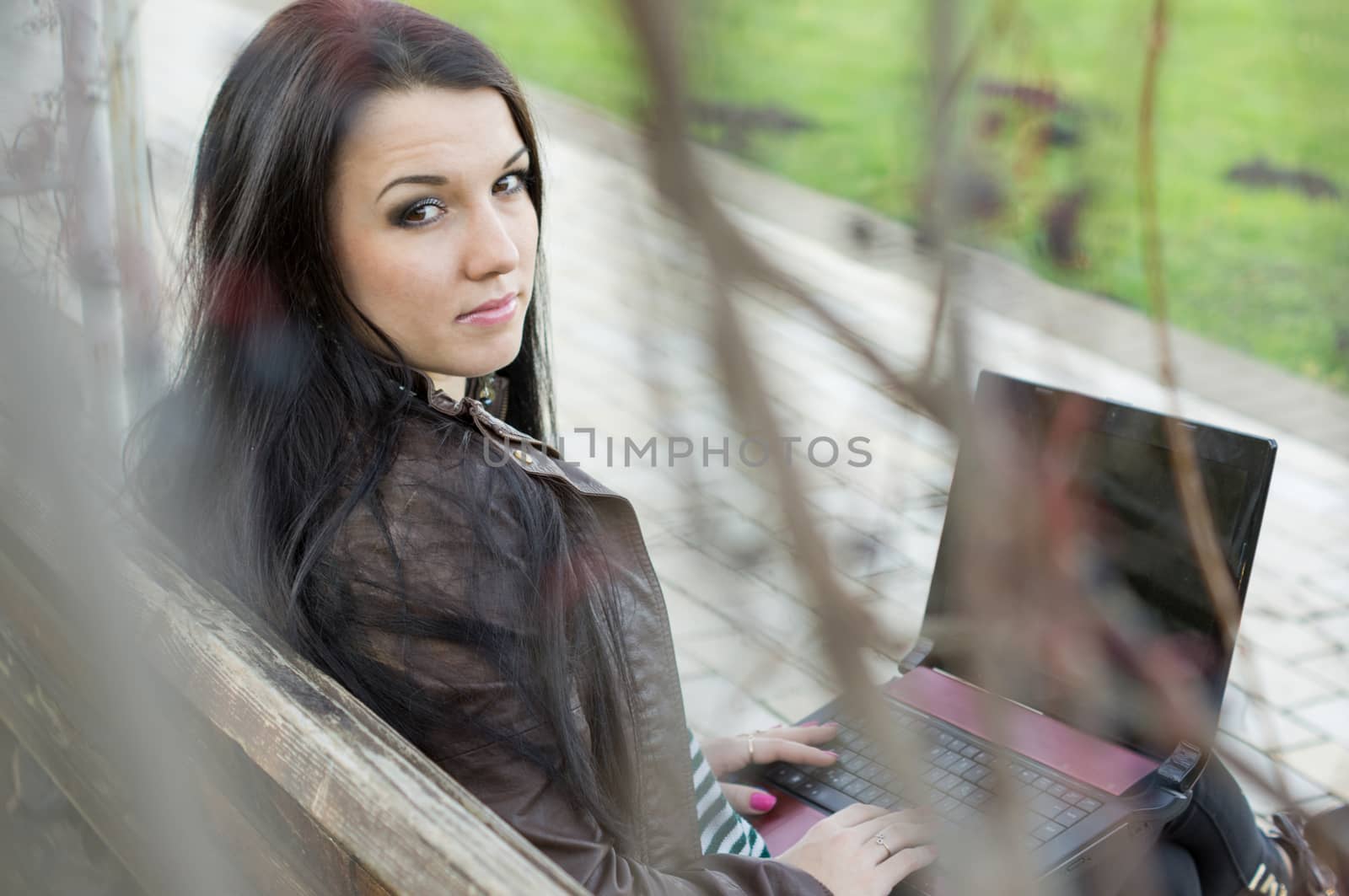 Young beautiful brunette student in profile sits on bench about university and works behind laptop.