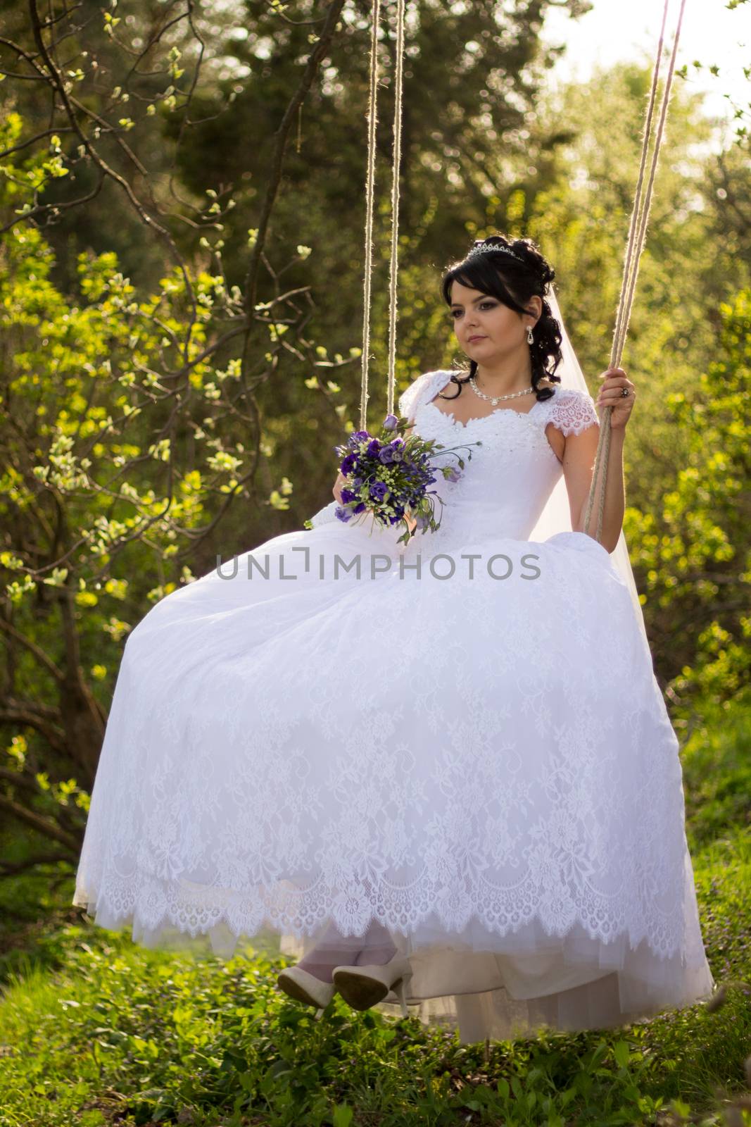 Portrait of a beautiful bride in white wedding dress sitting on swing outdoors