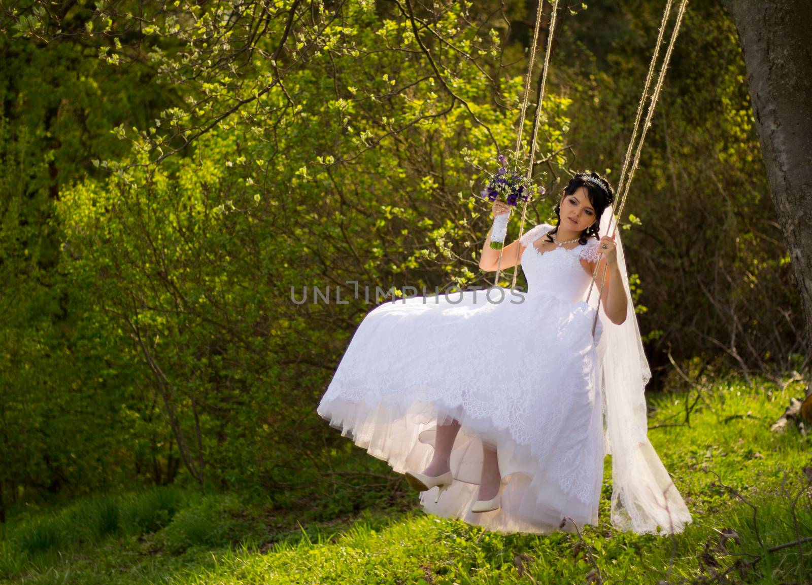 Portrait of a beautiful bride in white wedding dress sitting on swing outdoors