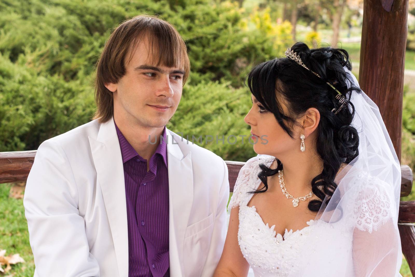 Couple in love bride and groom posing sitting on wooden bench in gazebo in their wedding day in summer.