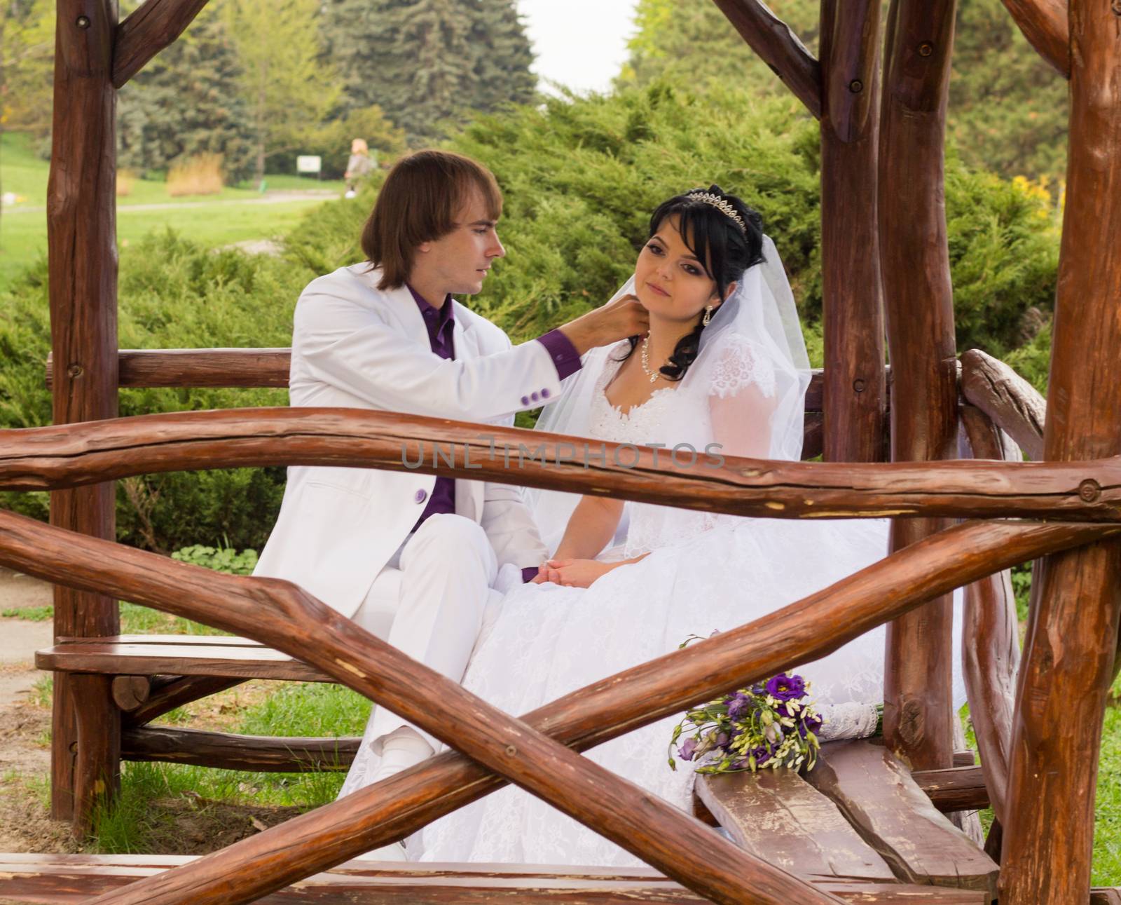 Couple in love bride and groom posing sitting on wooden bench in gazebo in their wedding day in summer.