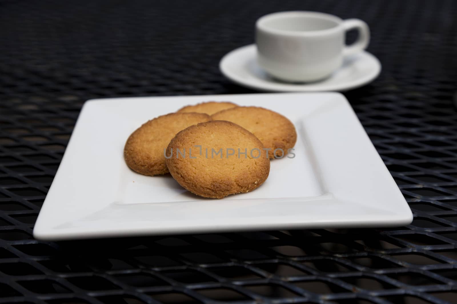 Selective focus on butter cookies on white plate with espresso visible in shallow depth of field.  Snack items on black metal mesh patio table.  