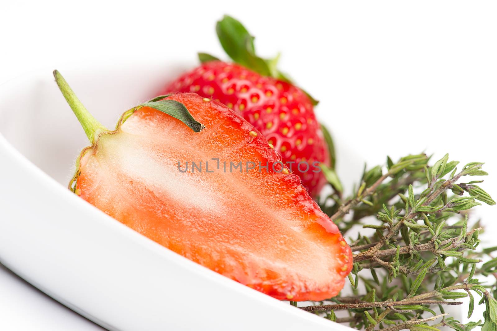 Halved fresh juicy ripe red strawberry showing the structure of the flesh or pulp in a white bowl with a sprig of fresh rosemary to be used as an aromatic flavoring, seasoning and cooking ingredient