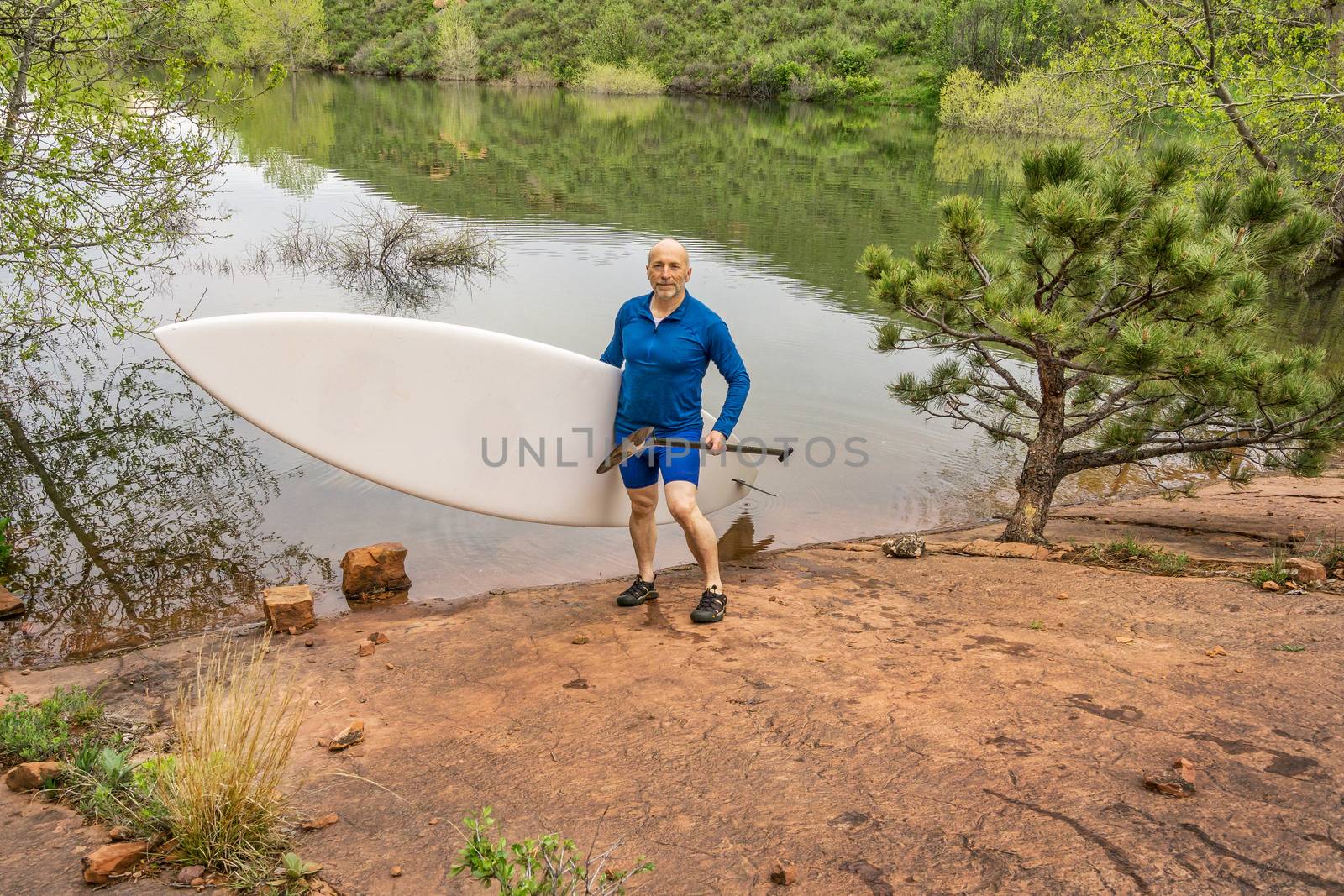senior paddler carrying paddleboard and paddle on a rocky shore of a lake - Horsetooth Reservoir, Fort Collins, Colorado