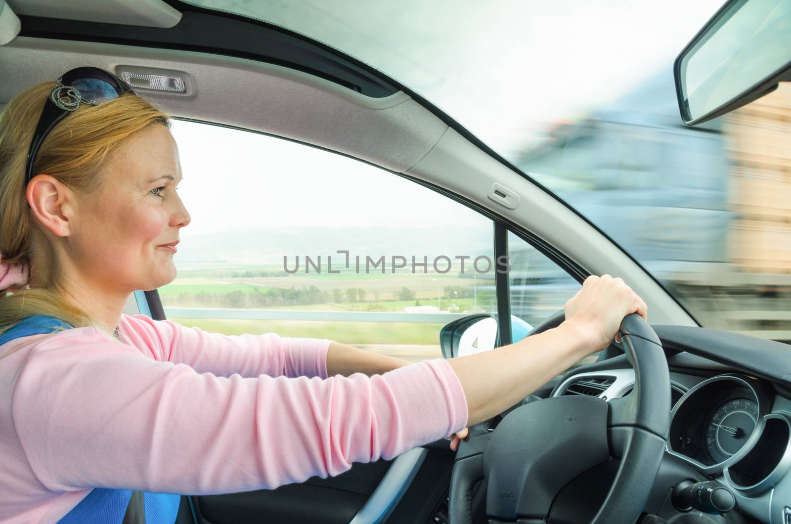 Attractive adult woman safe and carefully driving car on suburban road. Inside the auto photo with high speed oncoming lorry truck blurred in motion.