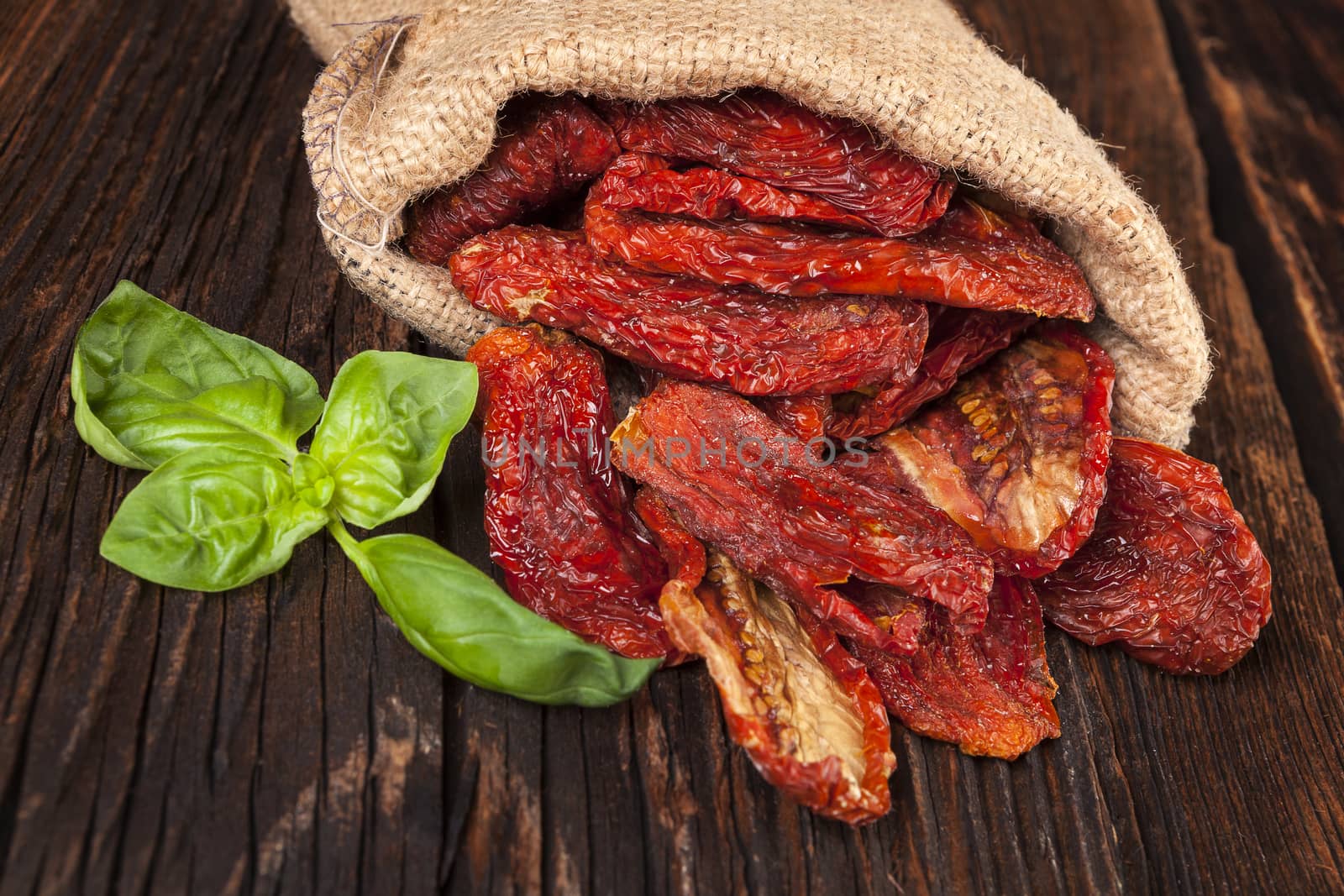 Delicious dried tomatoes with fresh basil leaves in burlap bag on brown wooden vintage textured background. Traditional mediterranean kitchen.