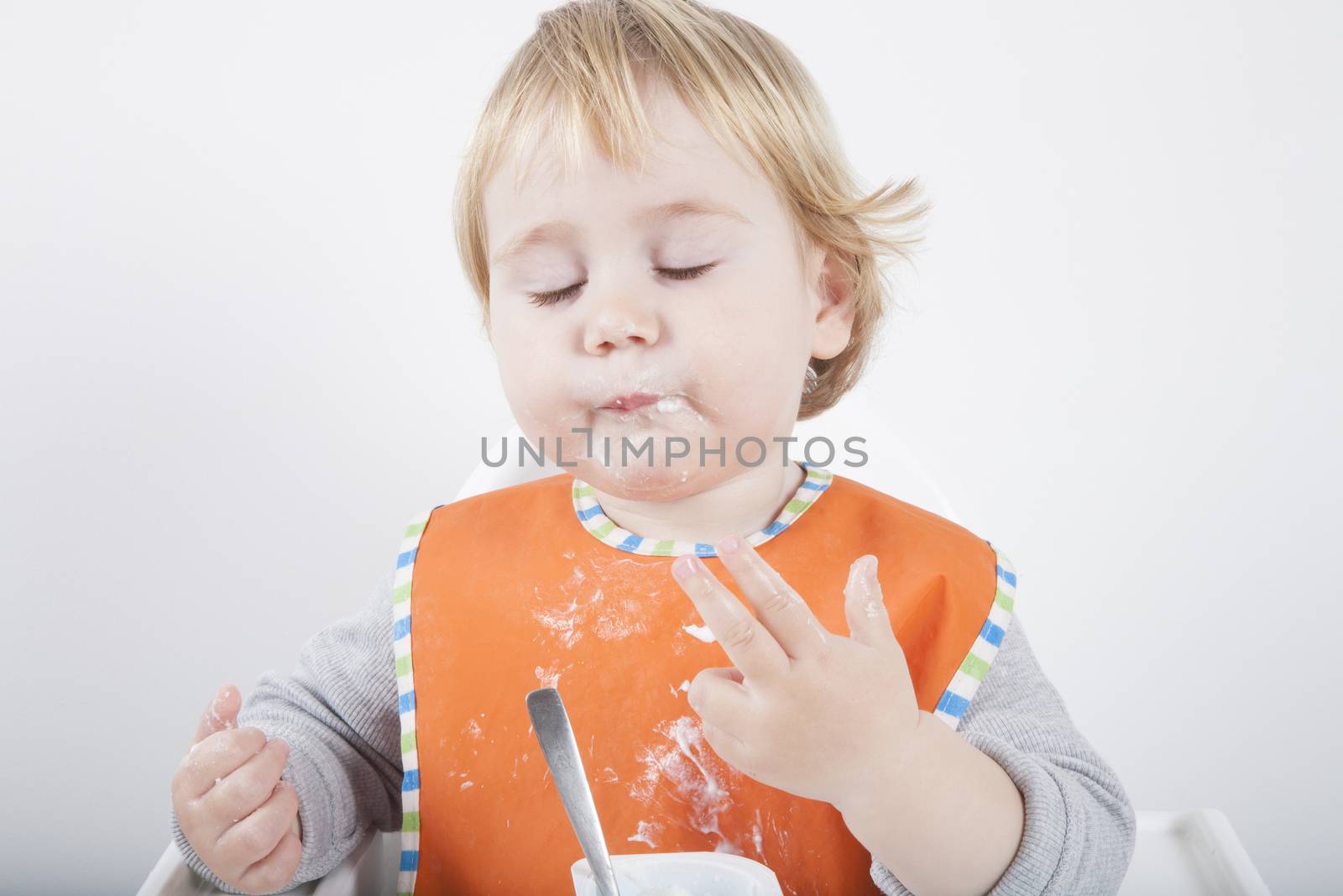 blonde caucasian baby seventeen month age orange bib grey sweater eating meal with her hand in white high chair