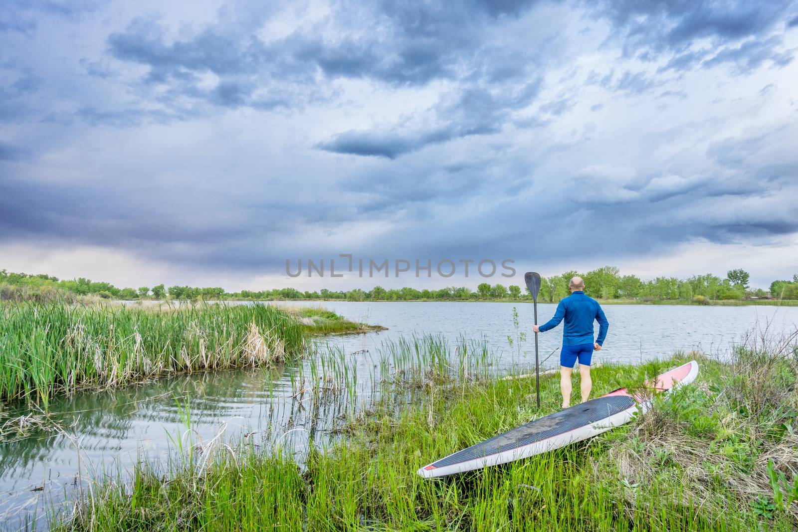 senior paddler with SUP paddleboar watching stormy sky by PixelsAway