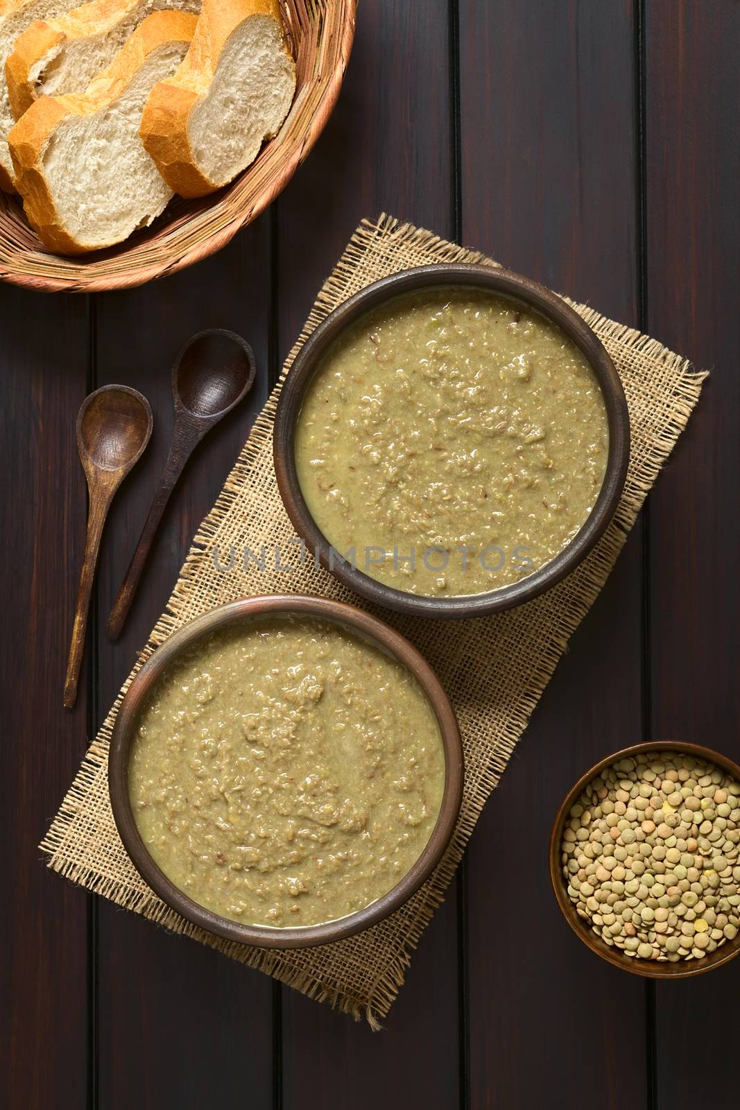 Cream of lentil soup in rustic bowls with wooden spoons, bread slices in basket and raw lentils in small bowl on the side, photographed overhead on dark wood with natural light
