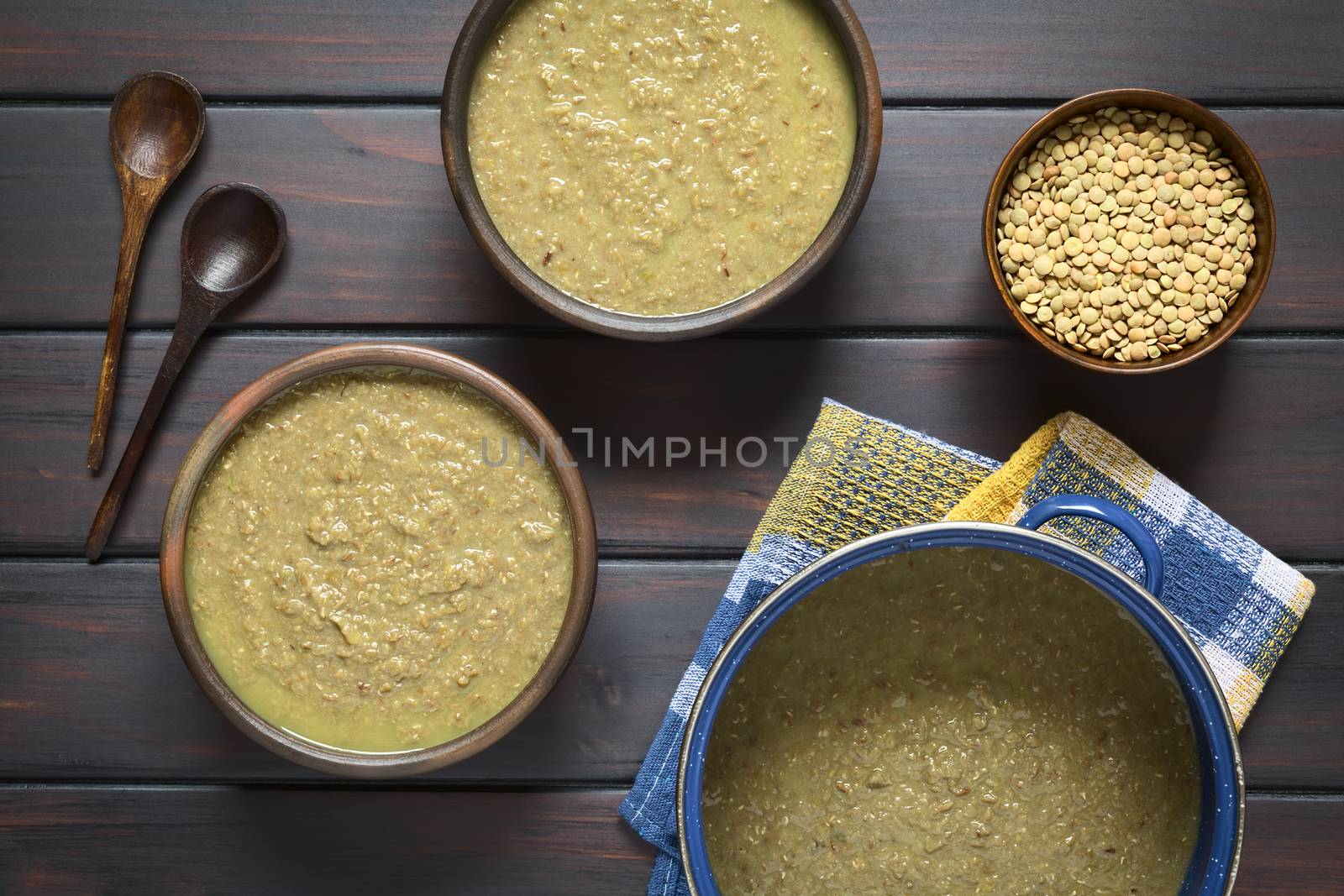 Cream of lentil soup in rustic bowls and cooking pot with wooden spoons and raw lentils in small bowl on the side, photographed overhead on dark wood with natural light