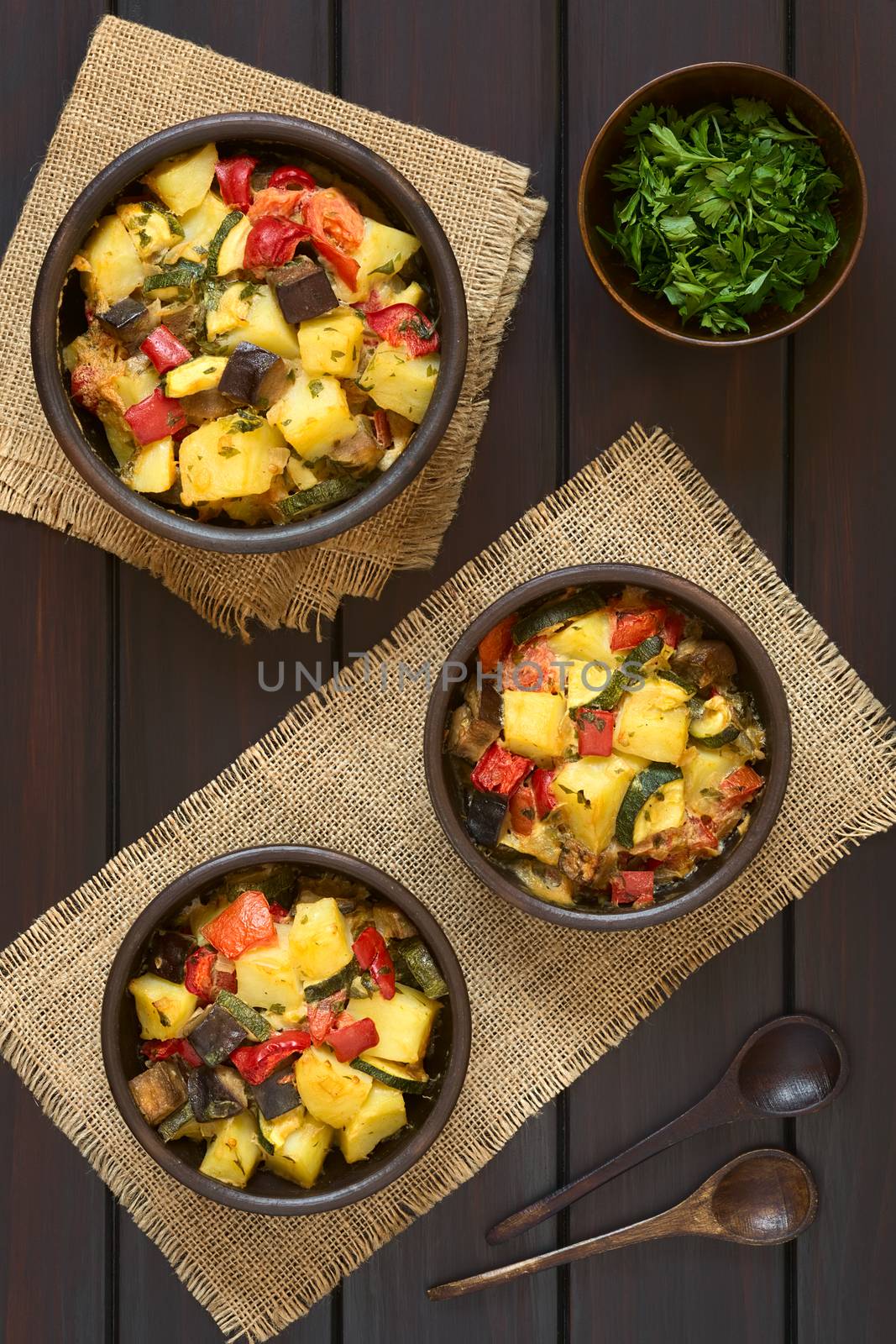 Baked potato, eggplant, zucchini and tomato casserole in rustic bowls, photographed overhead on dark wood with natural light