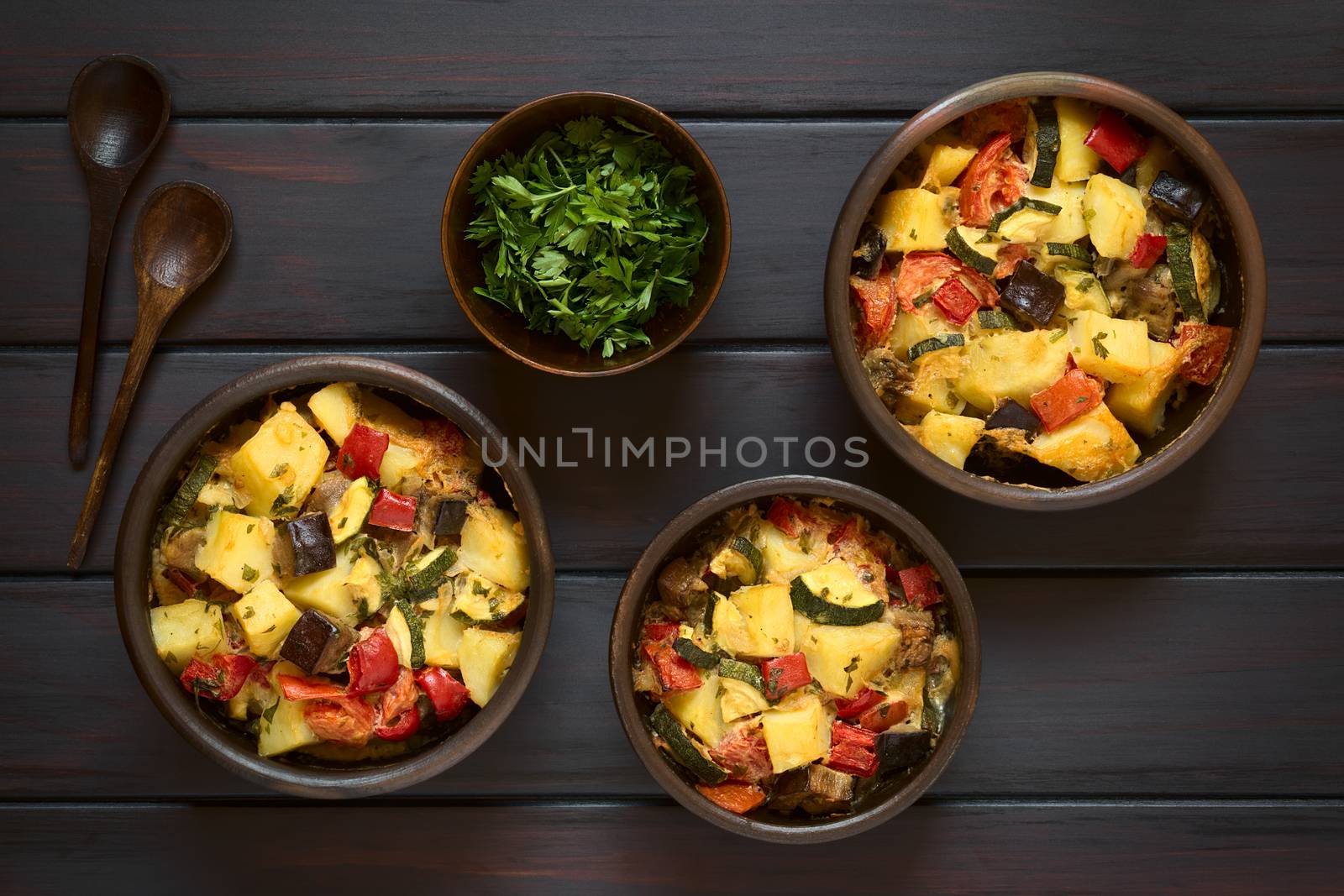 Baked potato, eggplant, zucchini and tomato casserole in rustic bowls, photographed overhead on dark wood with natural light