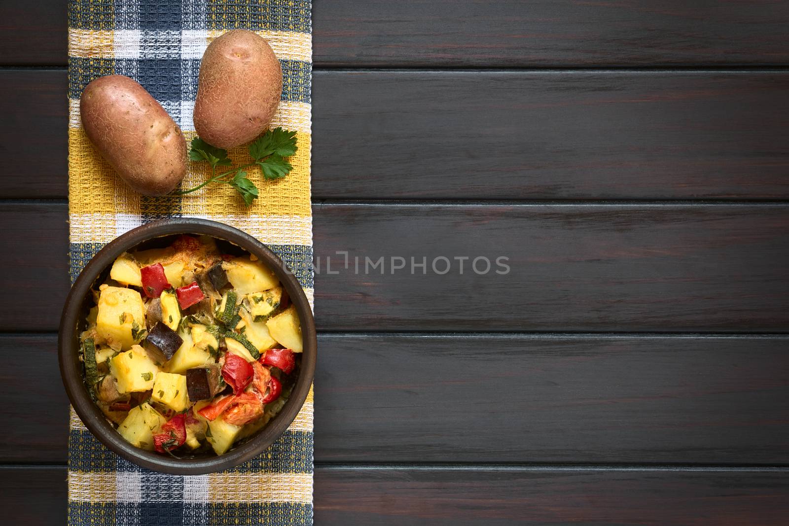 Baked potato, eggplant, zucchini and tomato casserole in rustic bowl, with raw potatoes and parsley leaf on kitchen towel, photographed overhead on dark wood with natural light