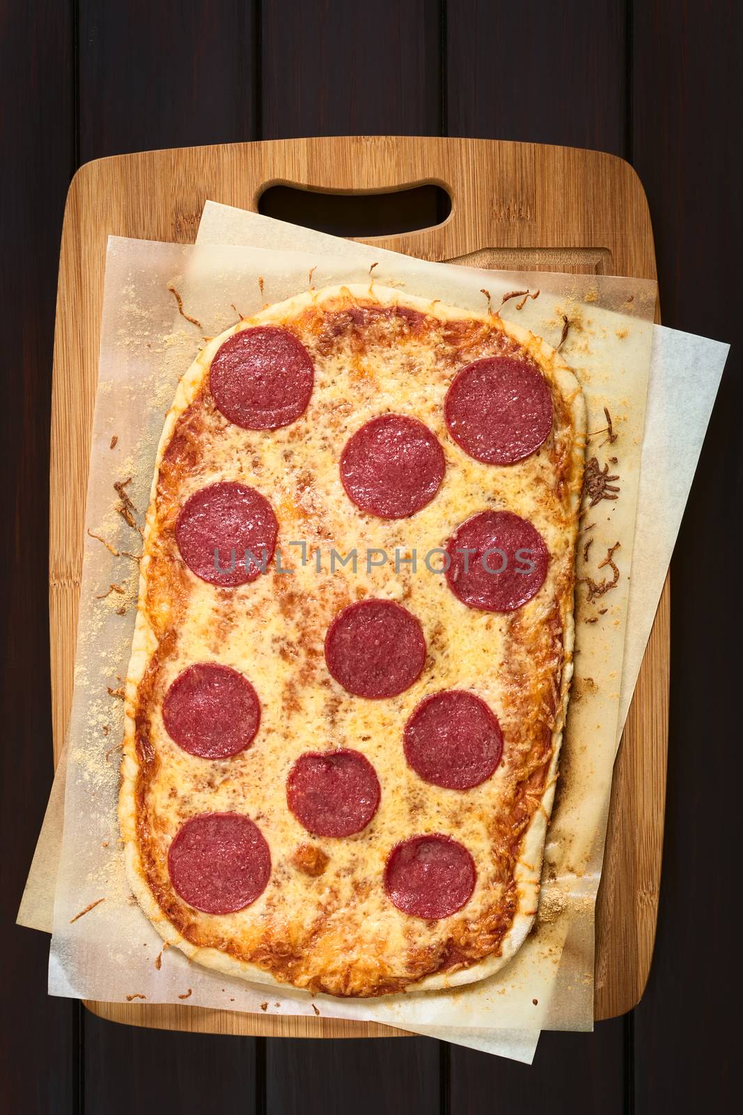 Homemade pepperoni or salami pizza on baking paper on wooden board, photographed overhead on dark wood with natural light