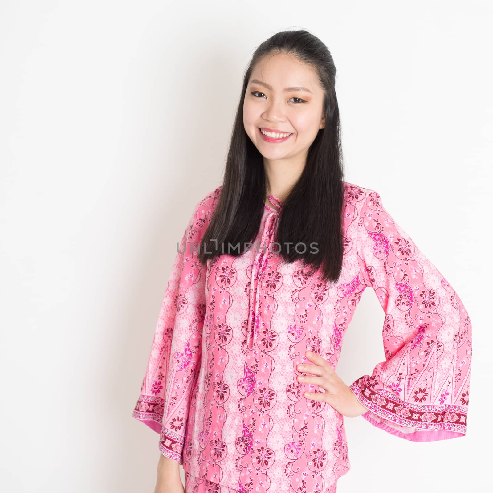 Portrait of happy Southeast Asian woman in pink batik dress standing on plain background.