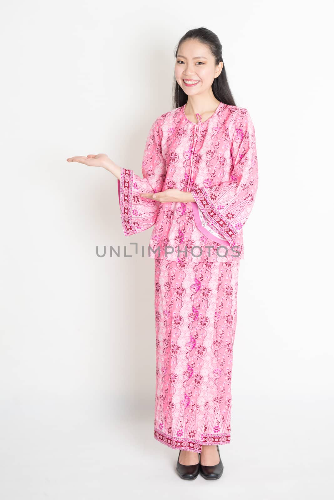 Full body portrait of happy Southeast Asian woman in pink batik dress hands showing something, standing on plain background.